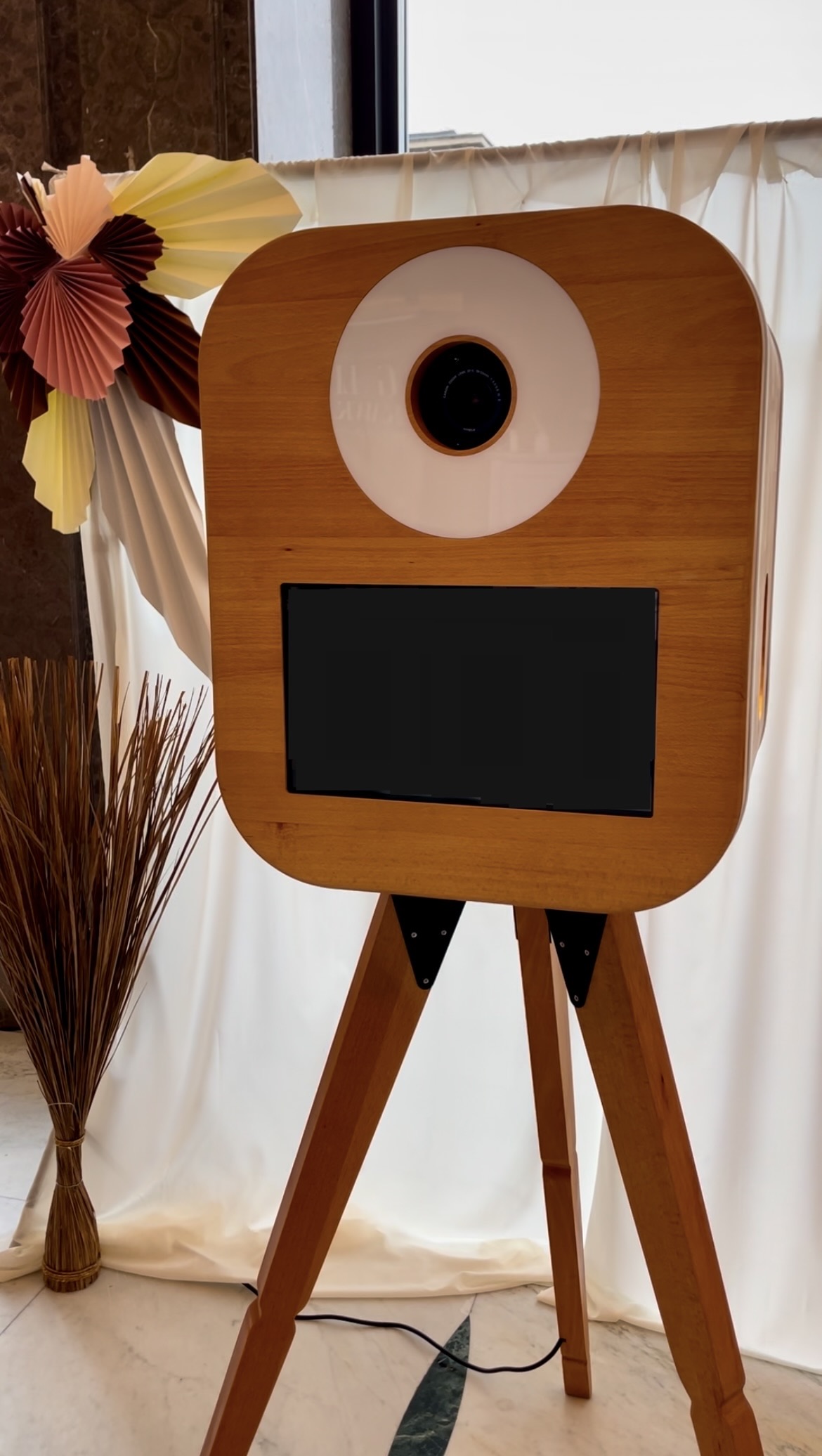 Drewniana fotobudka retro Harmonia na tle materiałowej ścianki z papierowymi kwiatami
